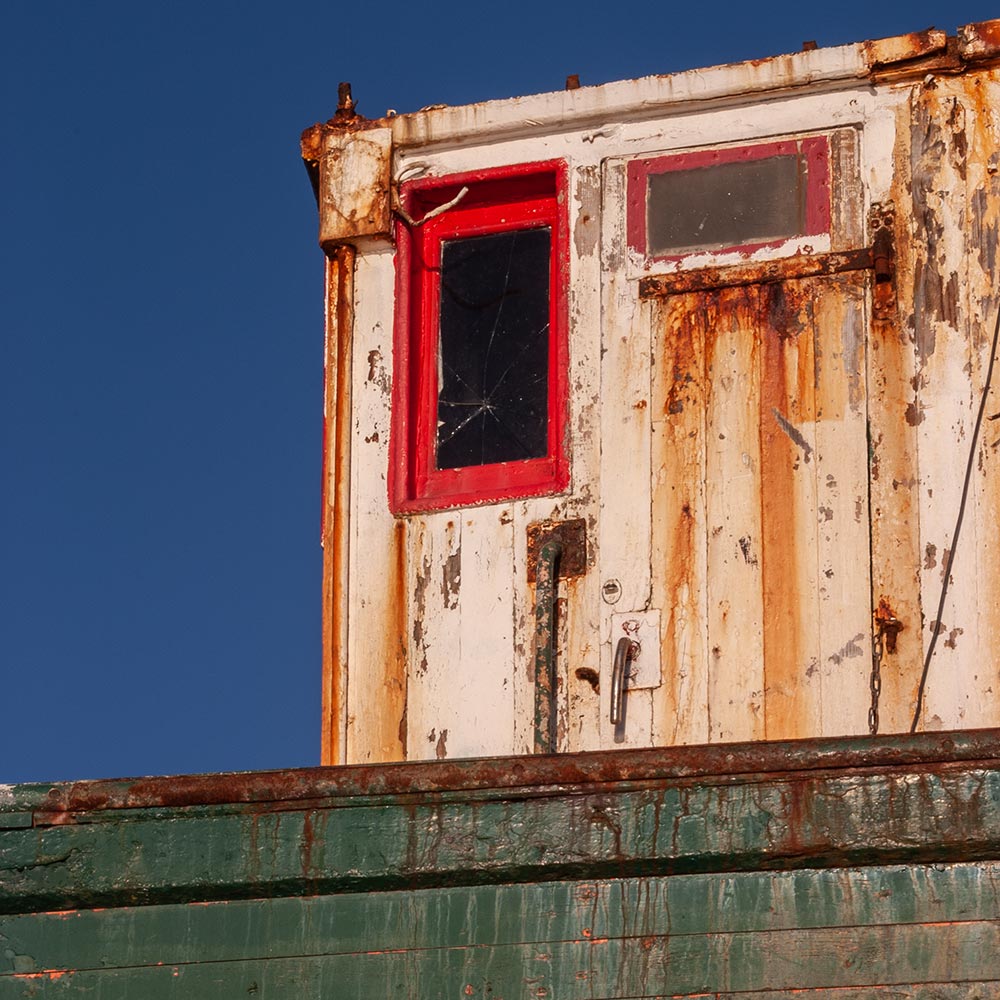 Camaret-sur_mer | a ships broken window with red painted frame on the rusting superstructure of a rotting hulk of a fishing boat with rust marks on the paint