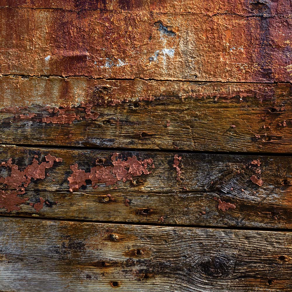 Camaret-sur_mer | The side of a burnt out rotting hulk of a fishing boat with peeling rusty red lead paint. Deeply textured worn panks of wood