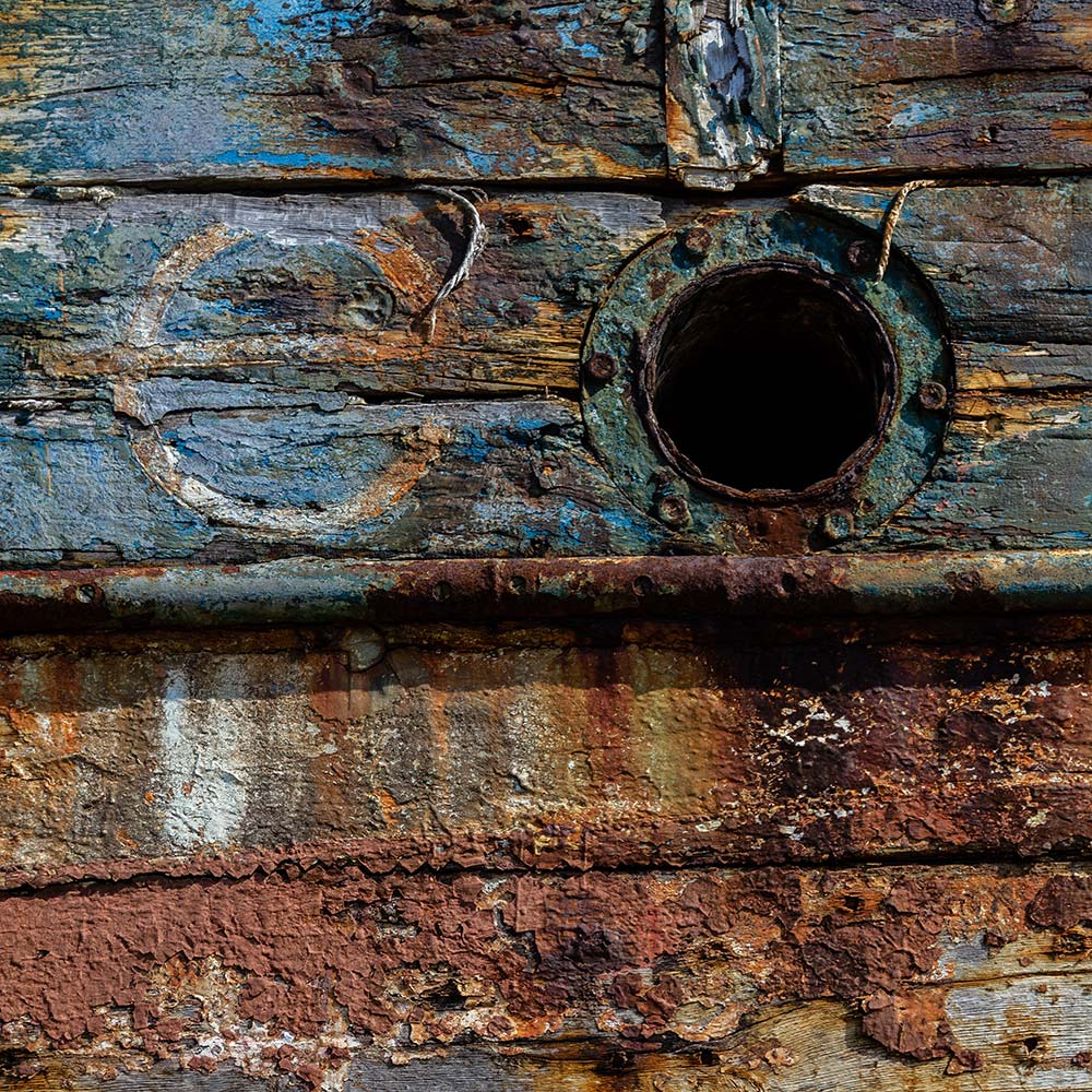 Camaret-sur_mer | The side of a burnt out rotting hulk of a fishing boat with peeling rusty white, blue and yellow paint with the design of a plimsoll line. Deeply textured worn panks of wood