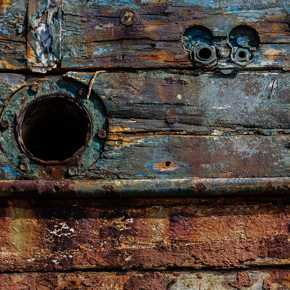 Camaret-sur_mer | The side of a burnt out rotting hulk of a fishing boat with waste pipes and peeling rusty white, blue and yellow painte. Deeply textured worn panks of wood with loose string caulking