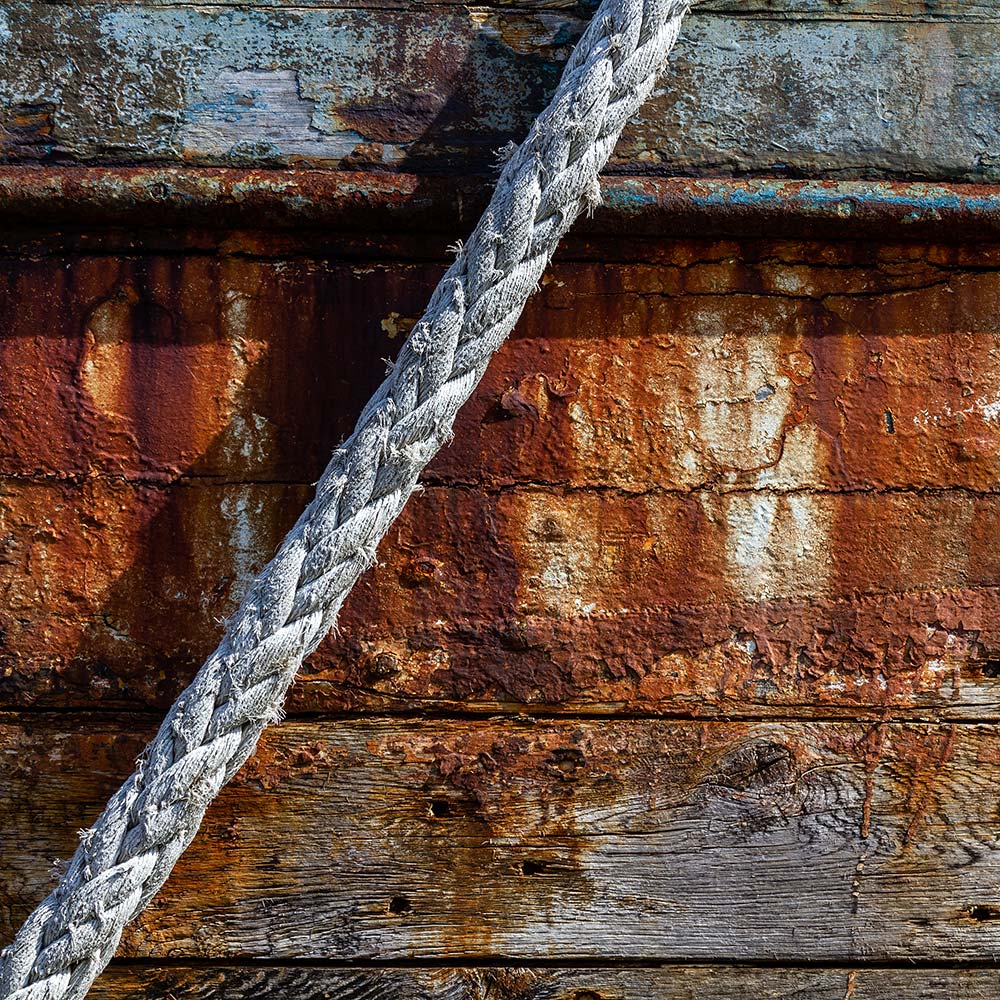Camaret-sur_mer | The side of a burnt out rotting hulk of a fishing boat with peeling rusty blue and ochre paint with old rope hauser. Deeply textured worn panks of wood with loose string caulking